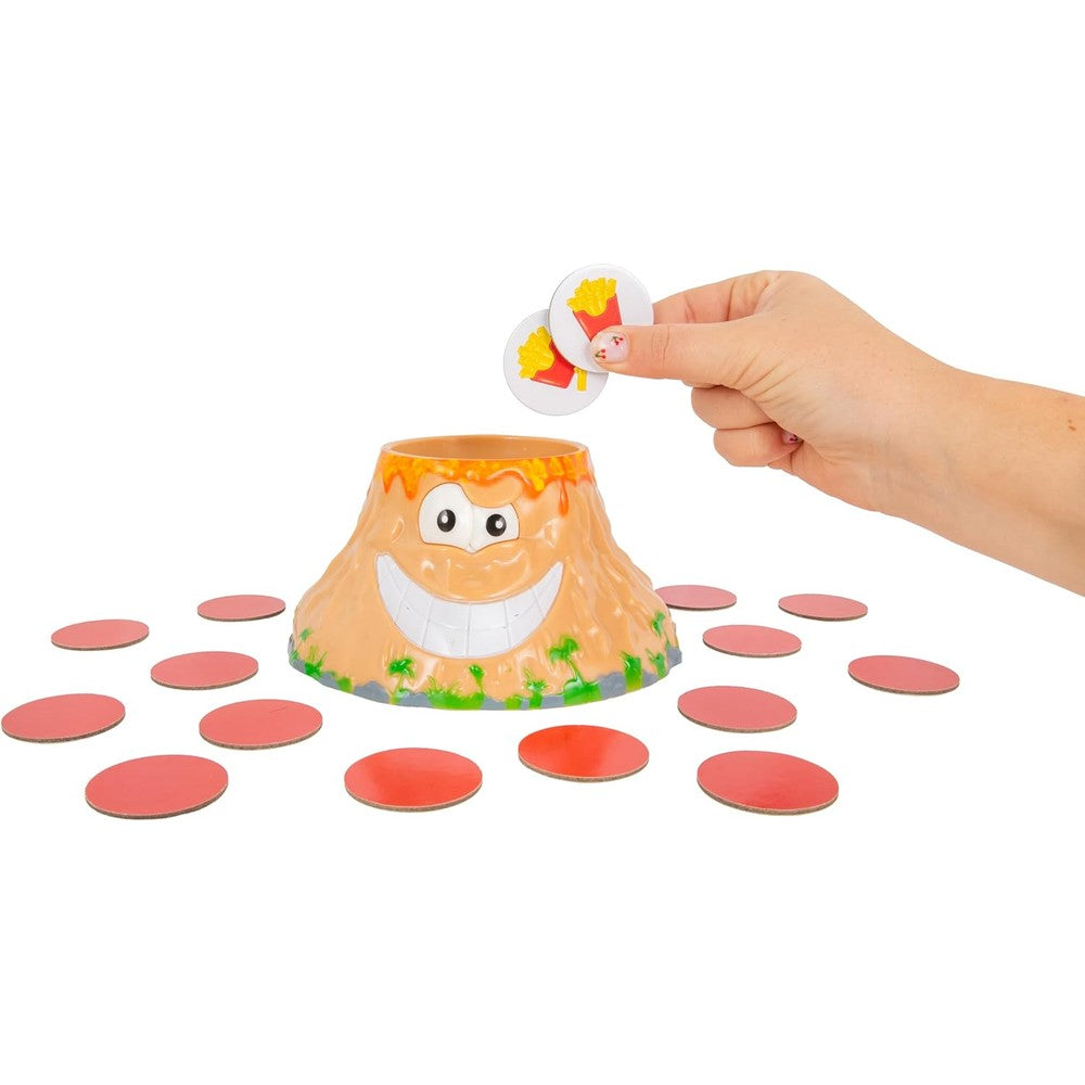 A hand holds two french fry tokens above the smiling lava-rimmed Volcanic Panic toy volcano surrounded by scattered red game pieces