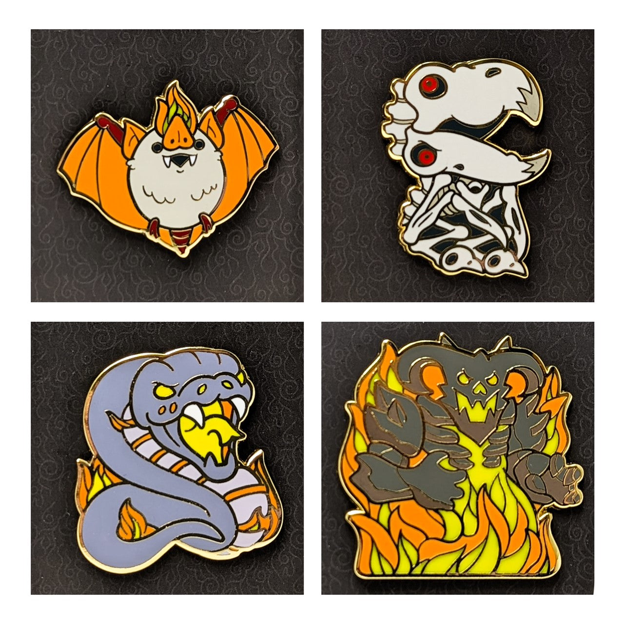 Four enamel pins: a cute bat with a flame, two stacked bird skulls, a coiled snake spitting fire, and a horned monster in roaring flames
