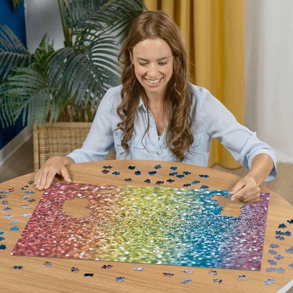 Smiling woman assembling a Ravensburger Glitter 1000-piece puzzle featuring a vibrant rainbow of sparkling colors on a wooden table