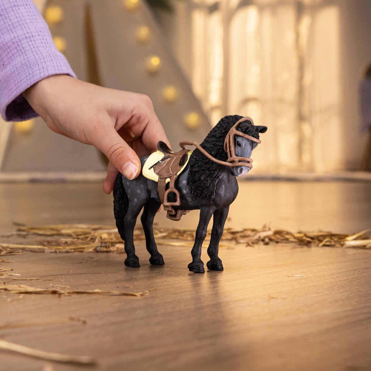 A child's hand holds a black Pura Raza Espanola Mare toy with a brown saddle and bridle on a wooden floor