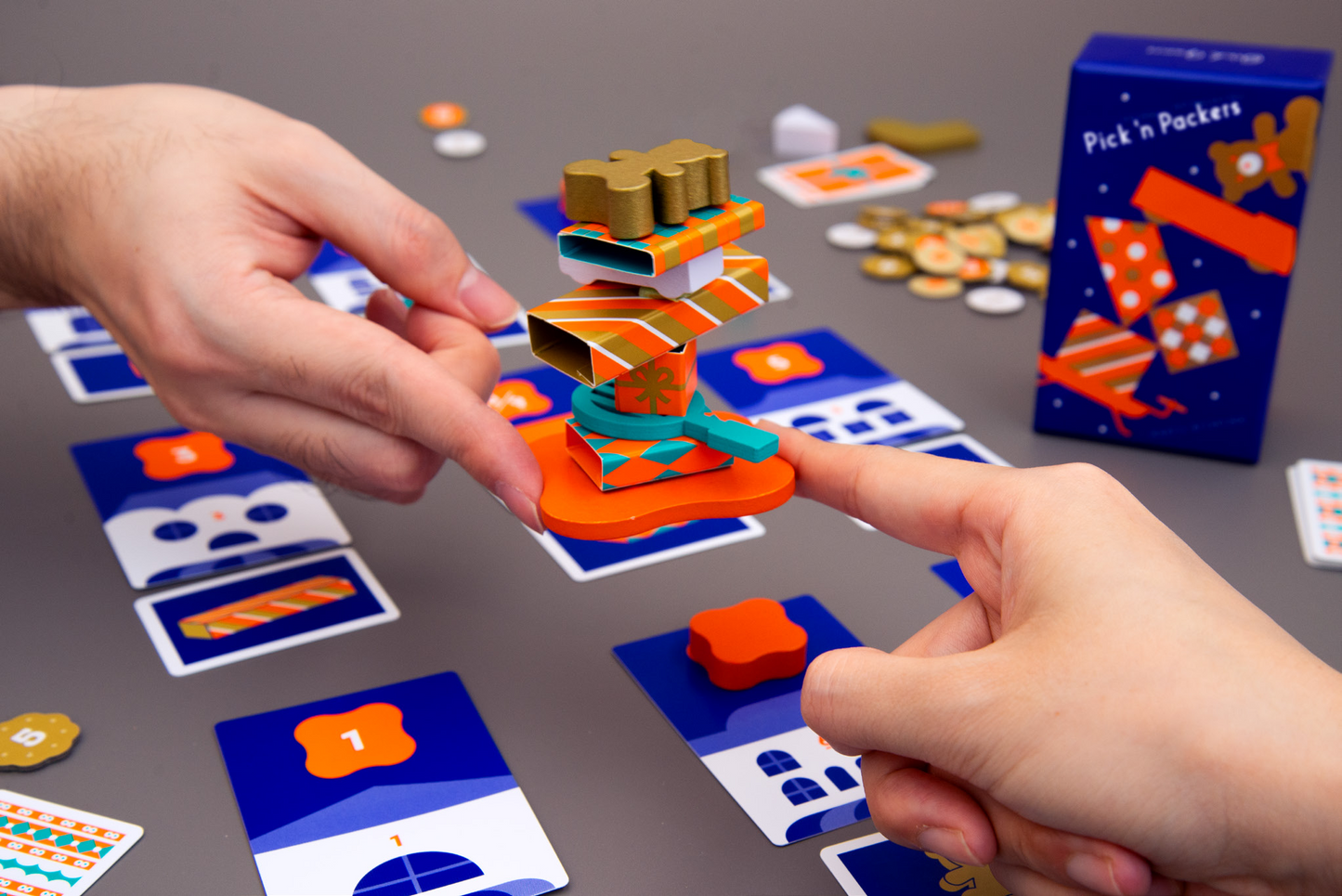 Hands balance stacked colorful boxes from the Pick 'n Packers game on an orange base, with game cards and tokens on a gray table