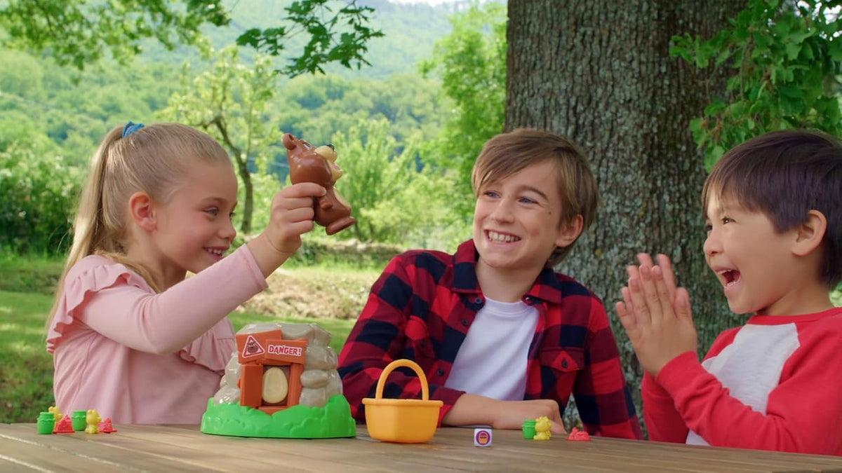 Three joyful children play an outdoor Beware of The Bear board game, a girl holds a toy bear while two boys laugh and clap