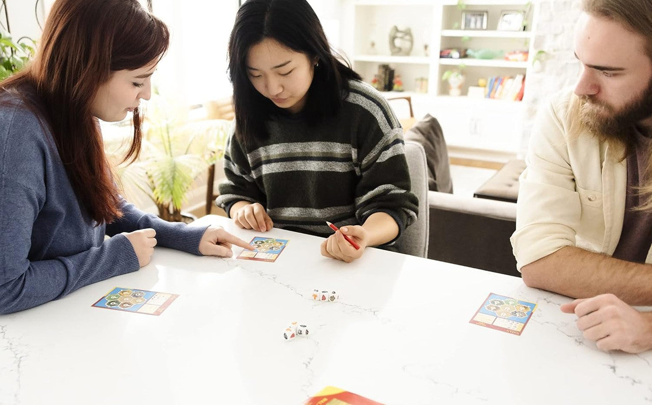 Two women and a man play a tabletop game at a white table. One woman points at a card; dice are on the table