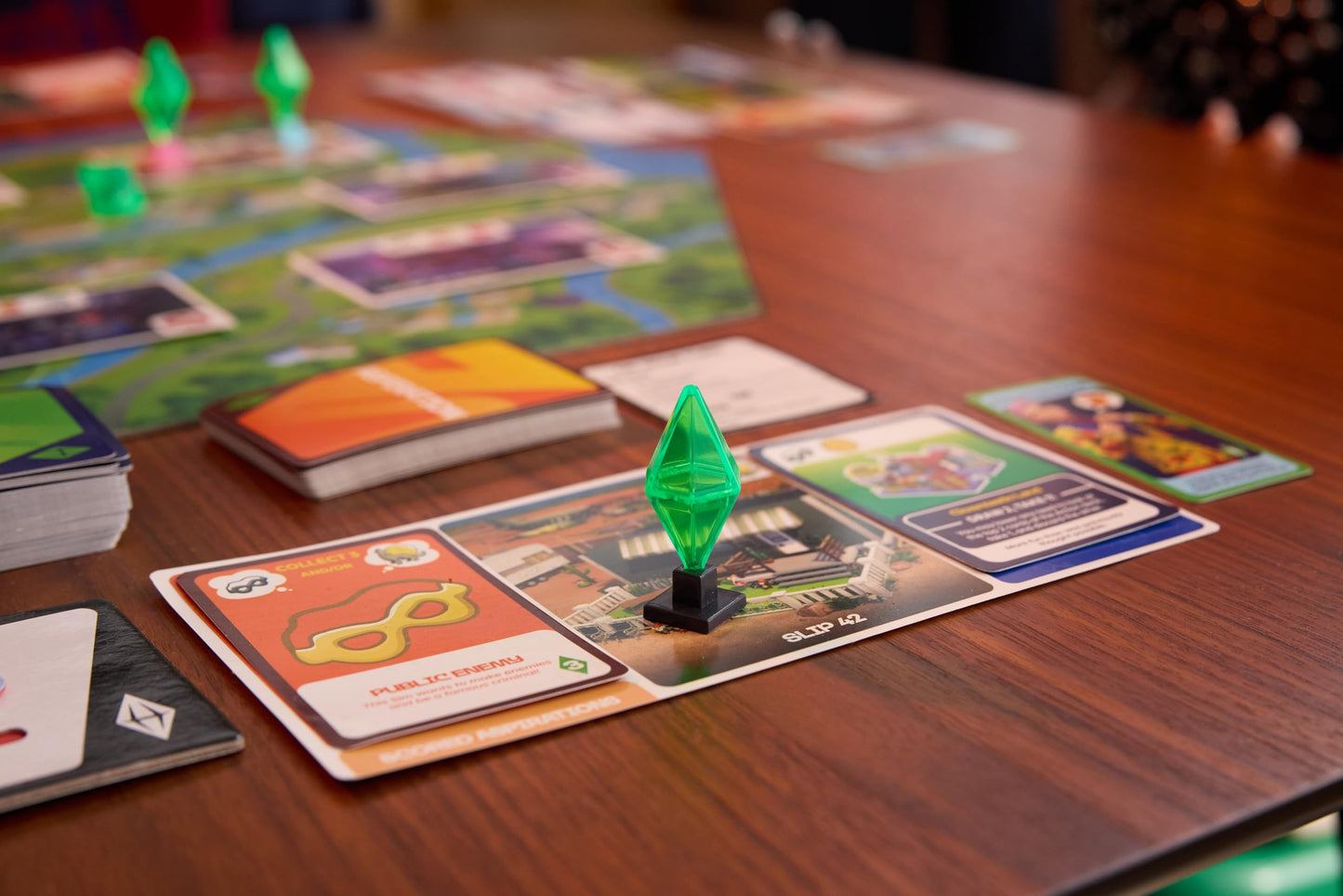 A centered view of The Sims board game in play on a wood table, highlighting a clear green Plumbob game piece placed on a character card next to stacks of orange and blue cards