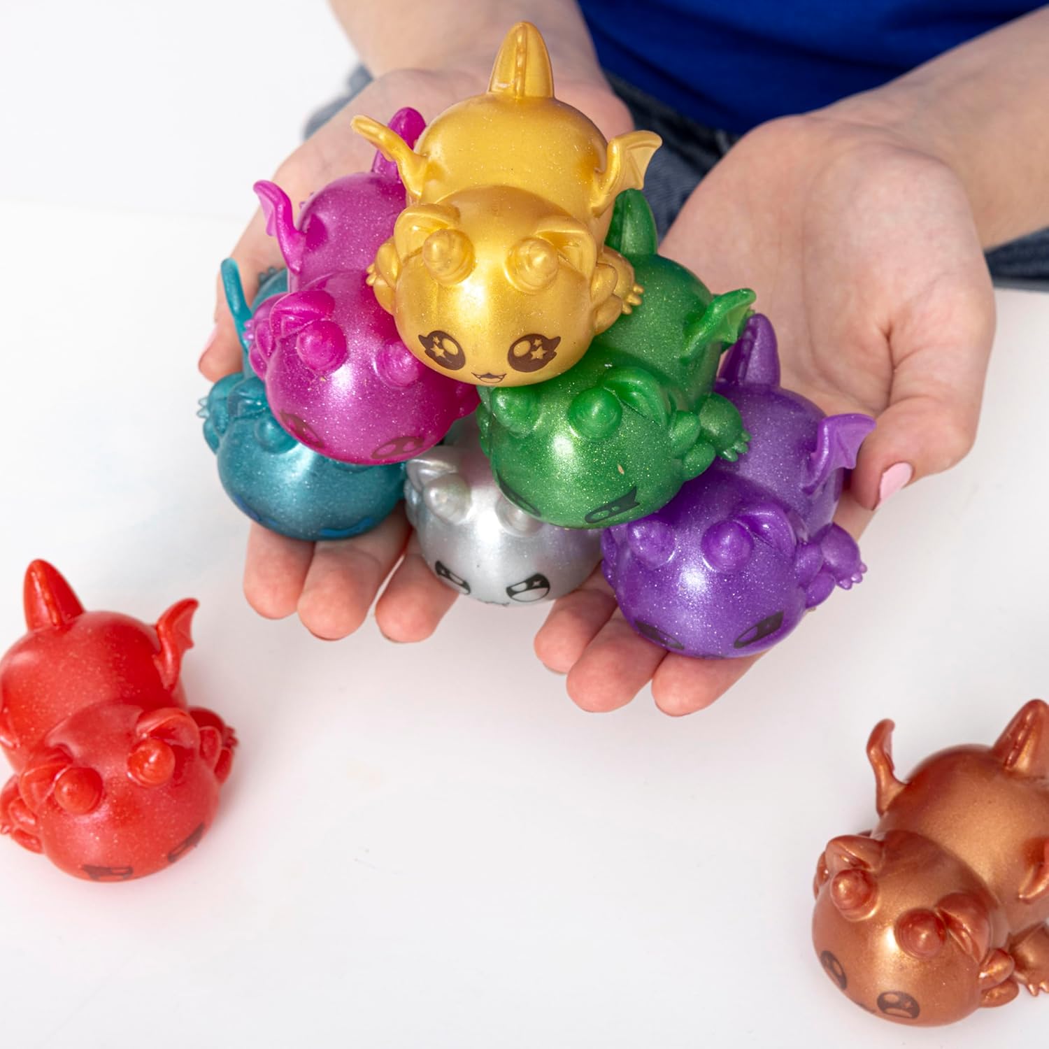 Hands hold a colorful stack of glittery monster figures: gold, green, purple, blue, pink, silver. Two more are on the white table