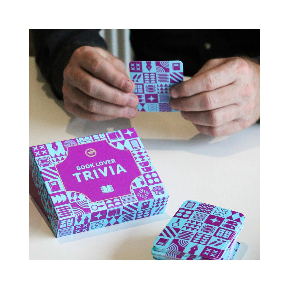 Hands hold a teal Book Lover Trivia card. The game box and card stack rest on a table