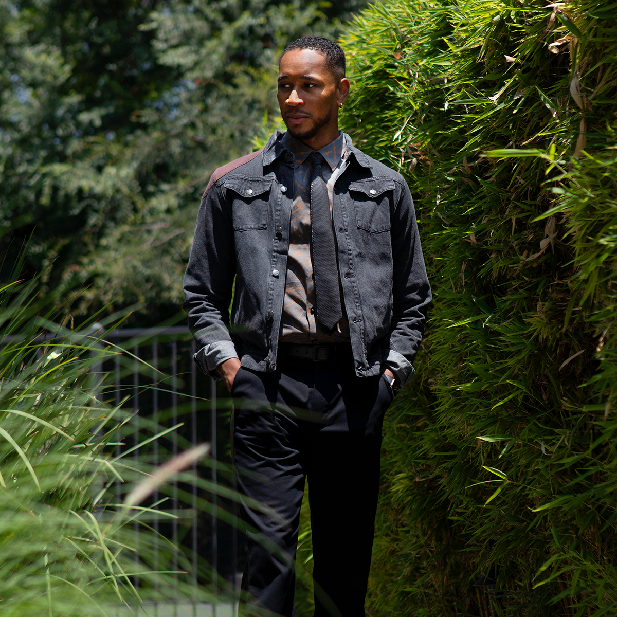 A Black man in a black denim jacket, patterned shirt, and tie walks with hands in pockets through a lush green outdoor space with a bamboo hedge