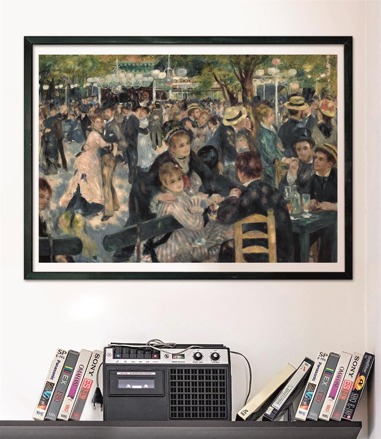 A framed painting of a bustling outdoor dance scene with many people hangs above a vintage cassette player and VHS tapes