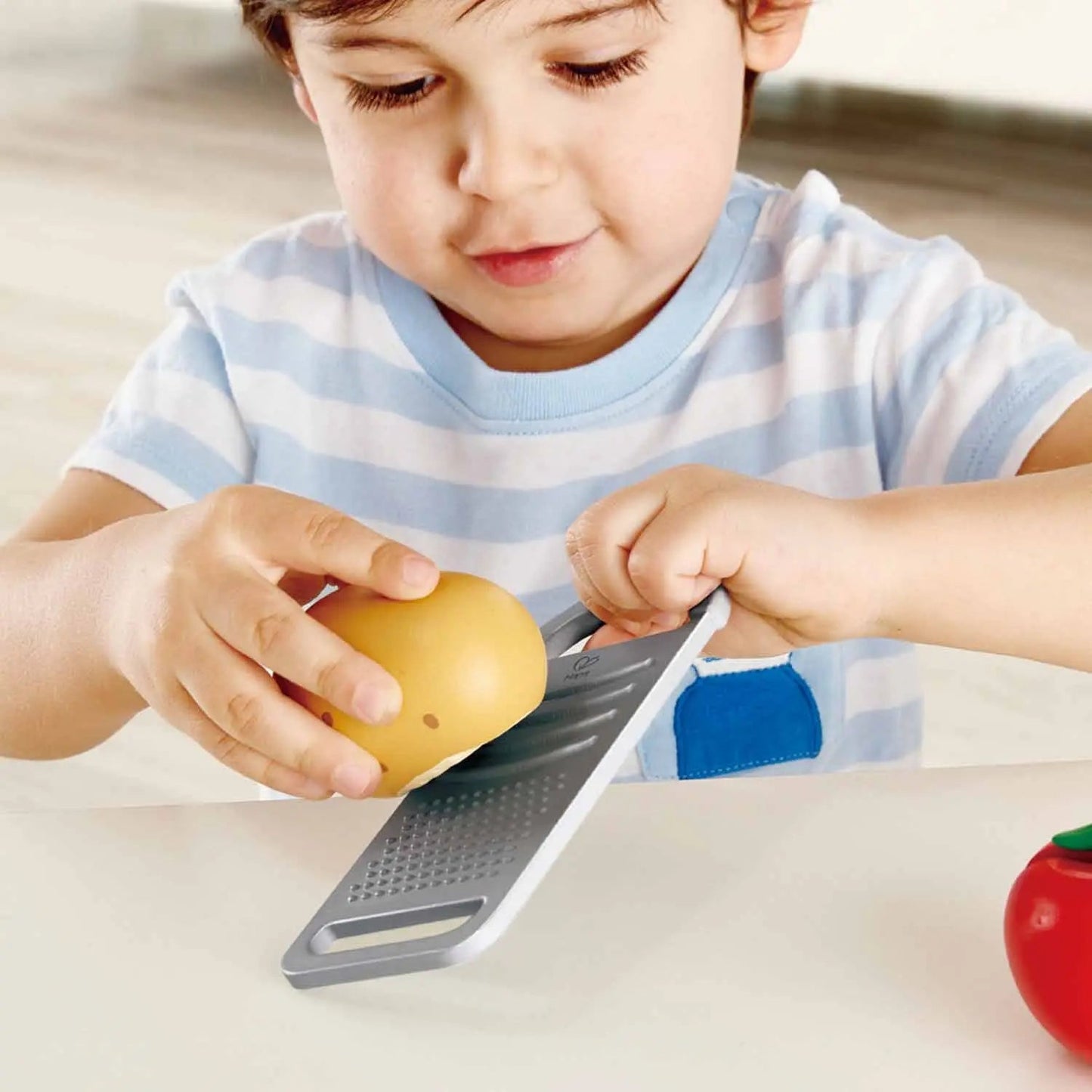 A young boy focuses on grating a yellow toy fruit with a toy grater.
