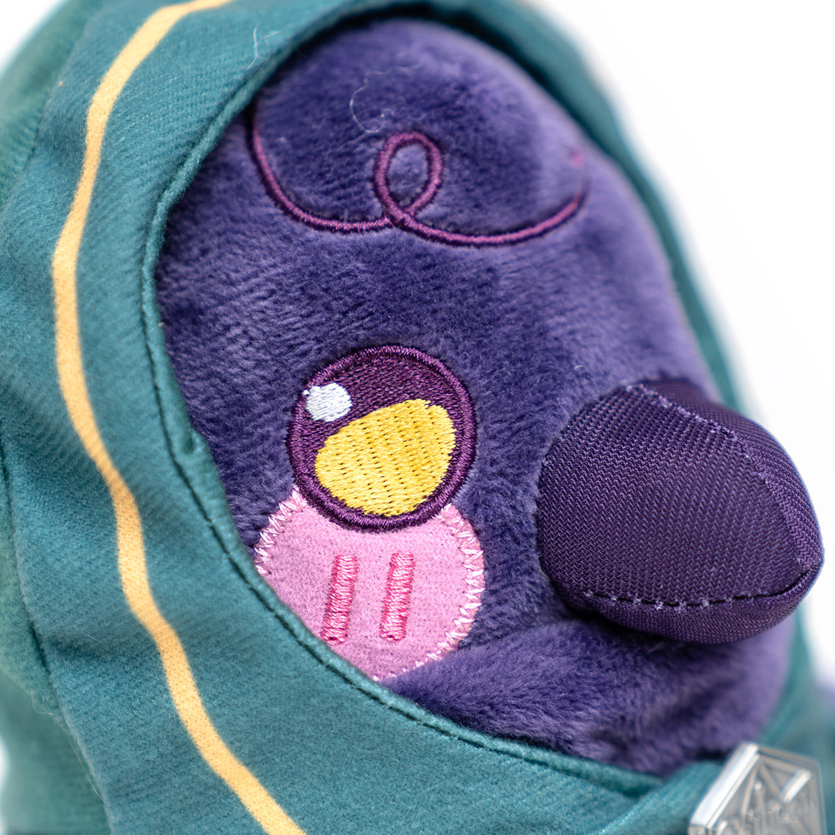 Close-up of a fluffy purple plush toy with one large yellow and white embroidered eye, pink lower face, and dark purple nose, wearing a striped teal hood