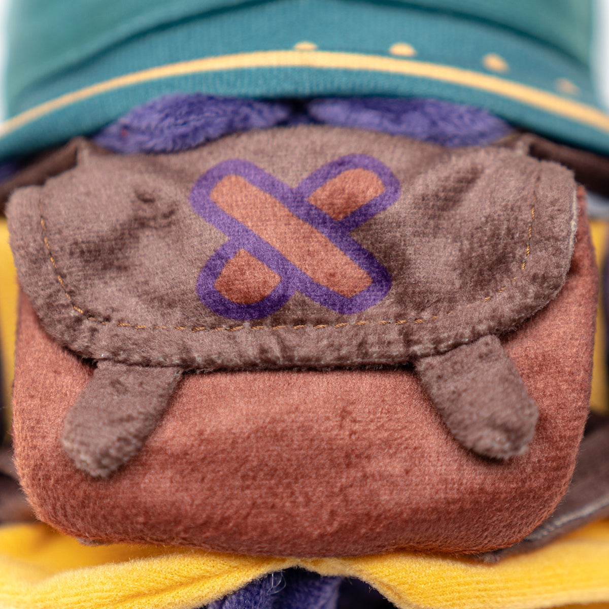 Plush character's brown satchel features a purple-outlined brown X-shaped symbol on its flap