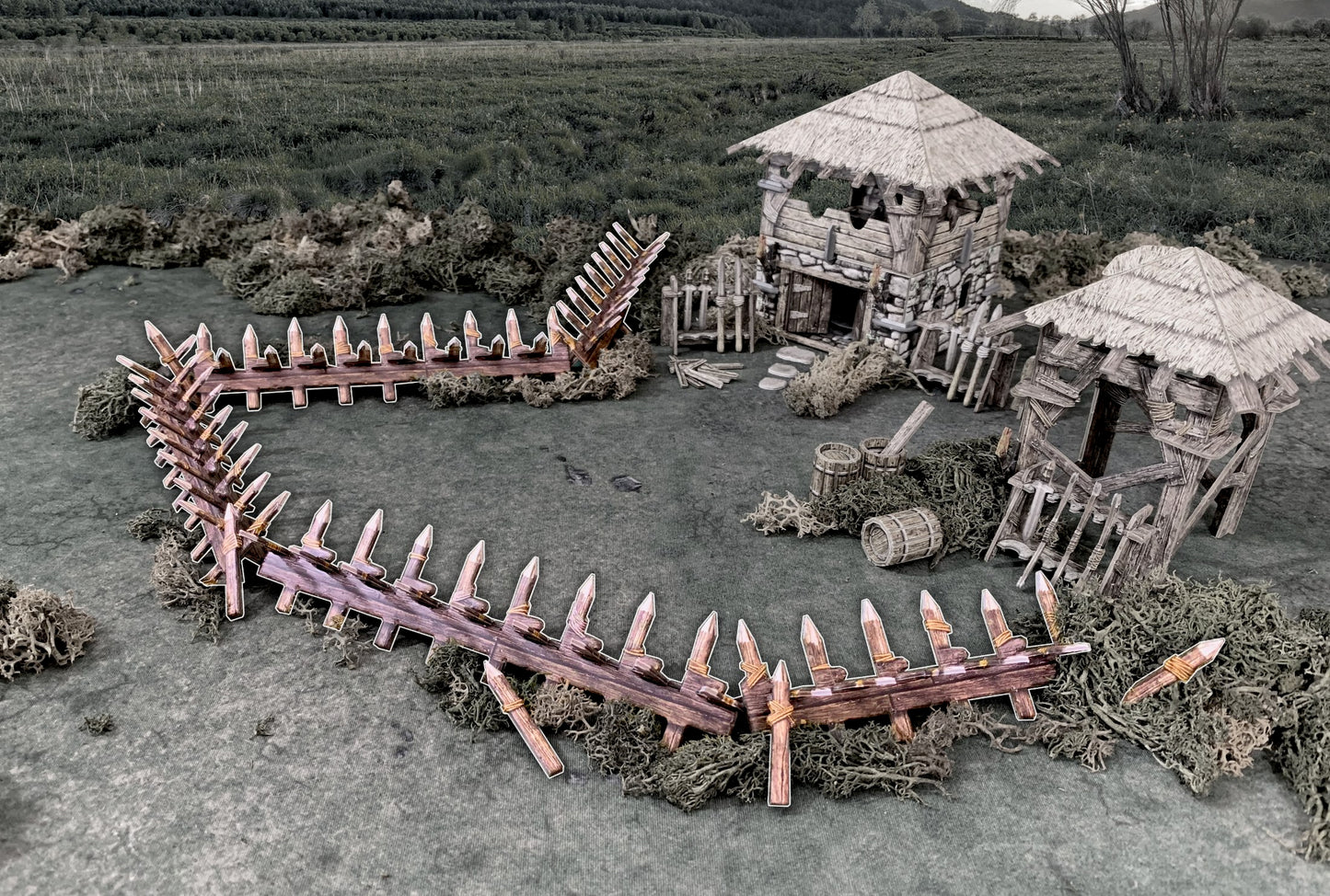 A miniature medieval village diorama with two thatched huts and a defensive wooden palisade on a grassy field.