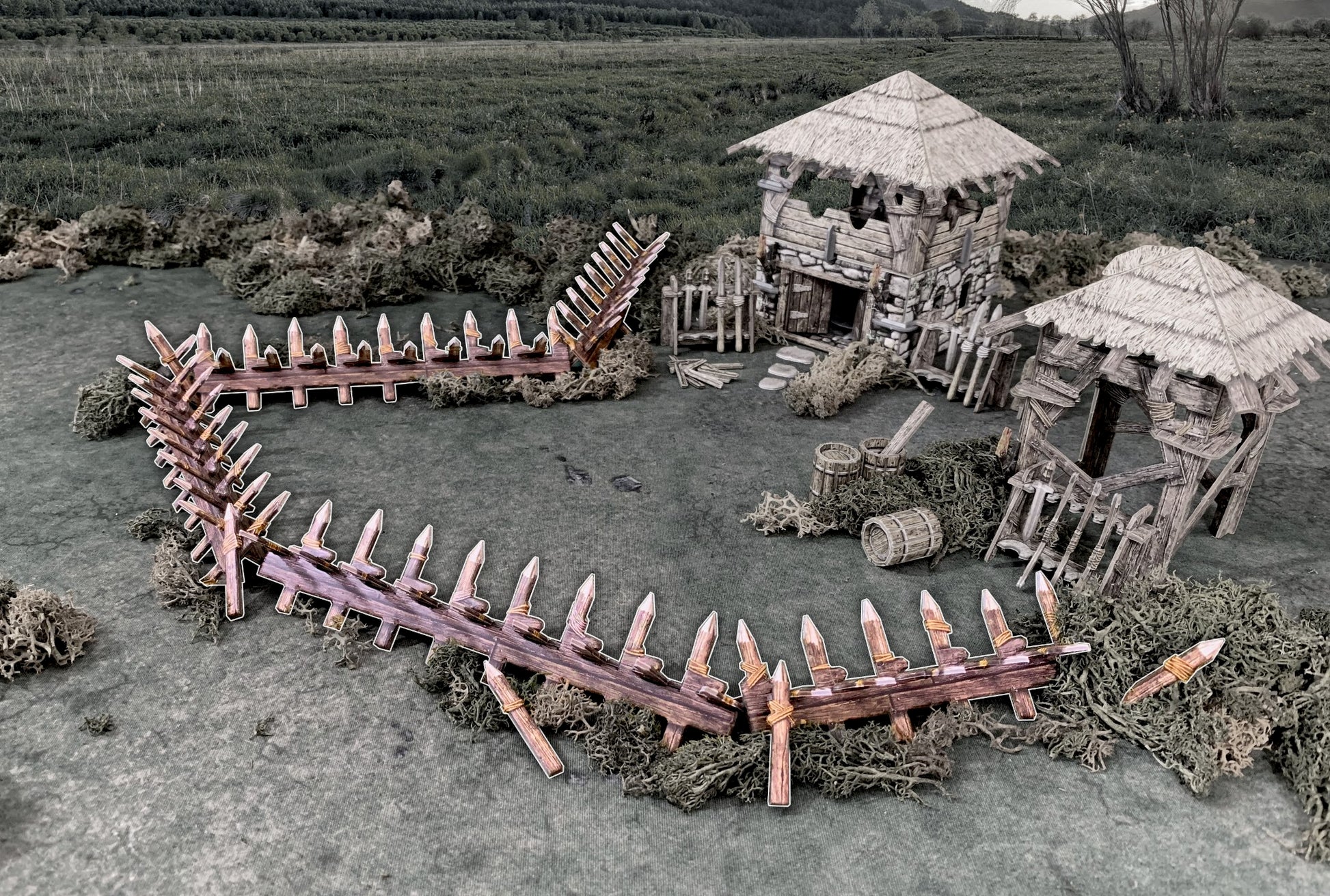 A miniature medieval village diorama with two thatched huts and a defensive wooden palisade on a grassy field.