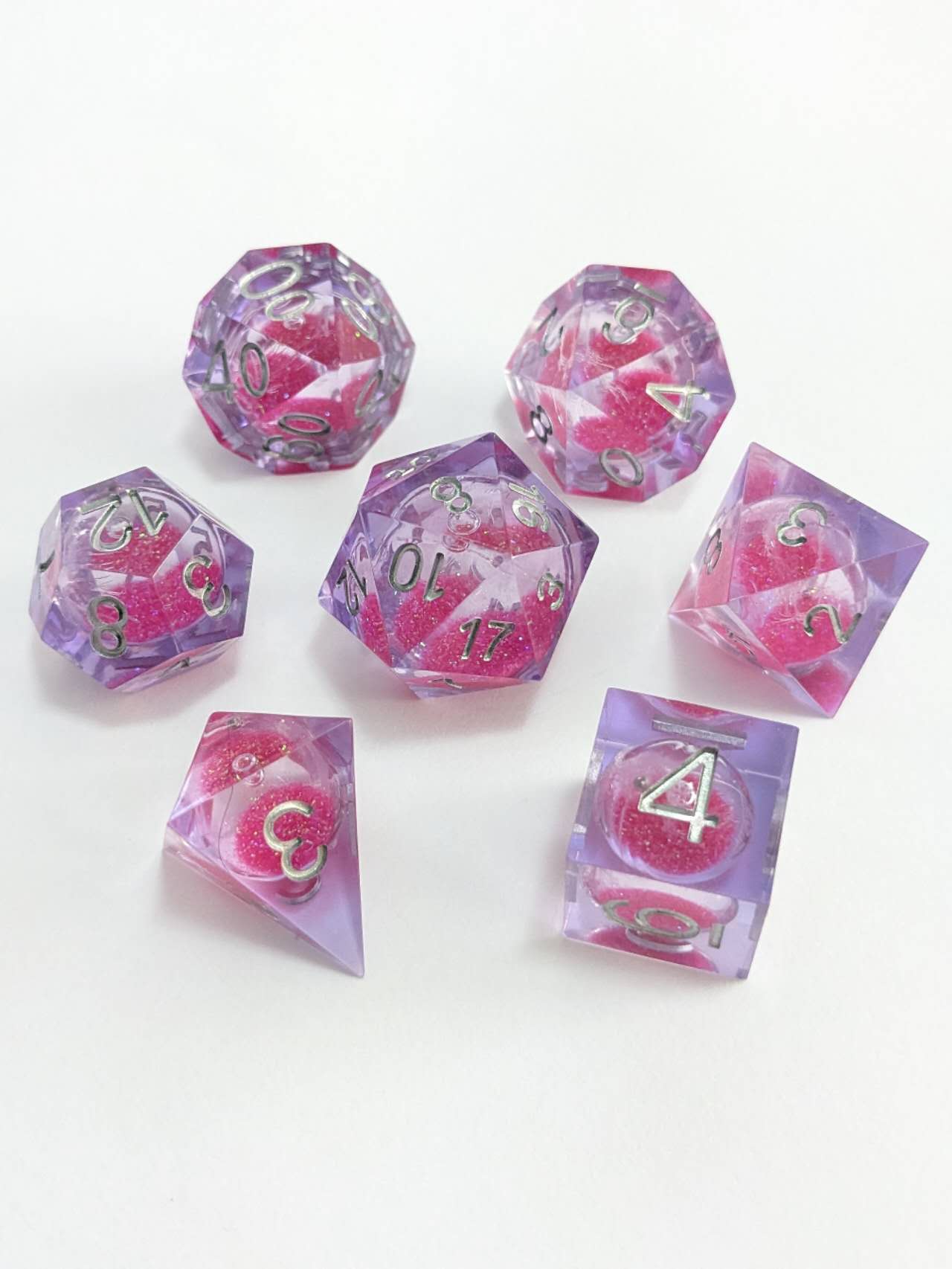 7 translucent purple polyhedral dice with pink glitter and silver numbers, arranged on a white surface