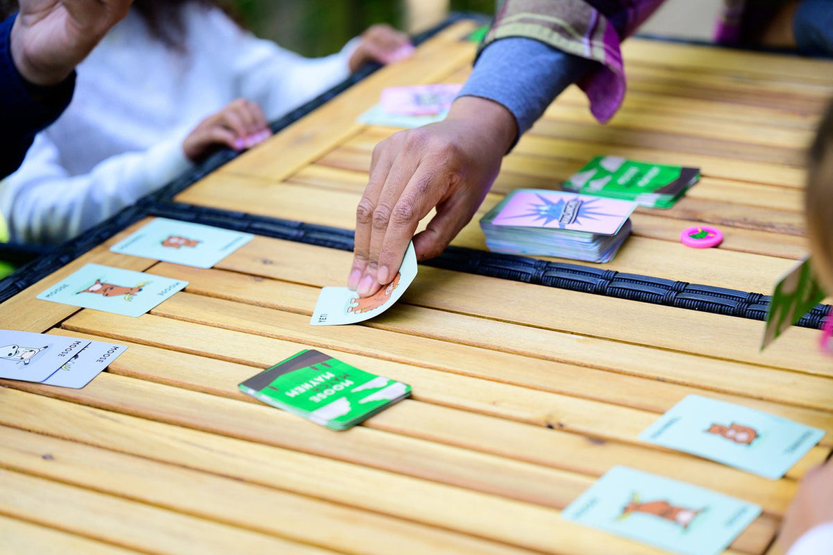 Hand placing a card during a game of Moose Mayhem on a wooden table, surrounded by other players