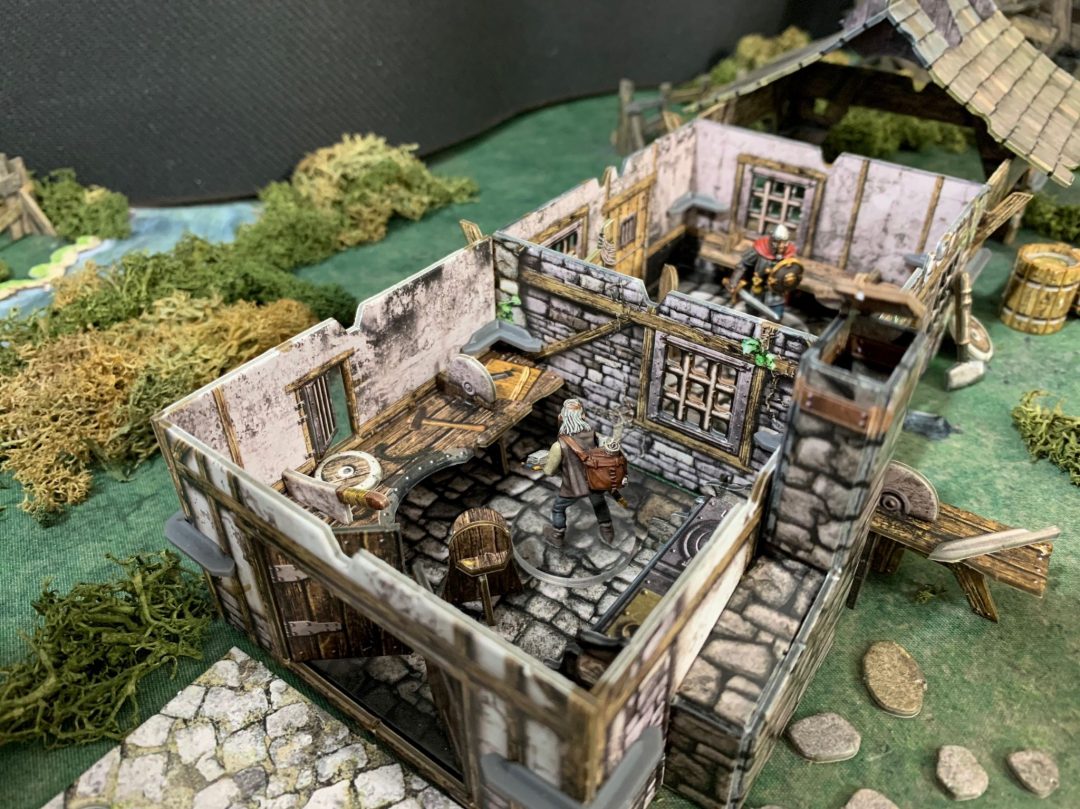 Detailed miniature diorama of a fantasy smithy, roof removed. A white-haired figure stands at a workbench, a warrior in the back.