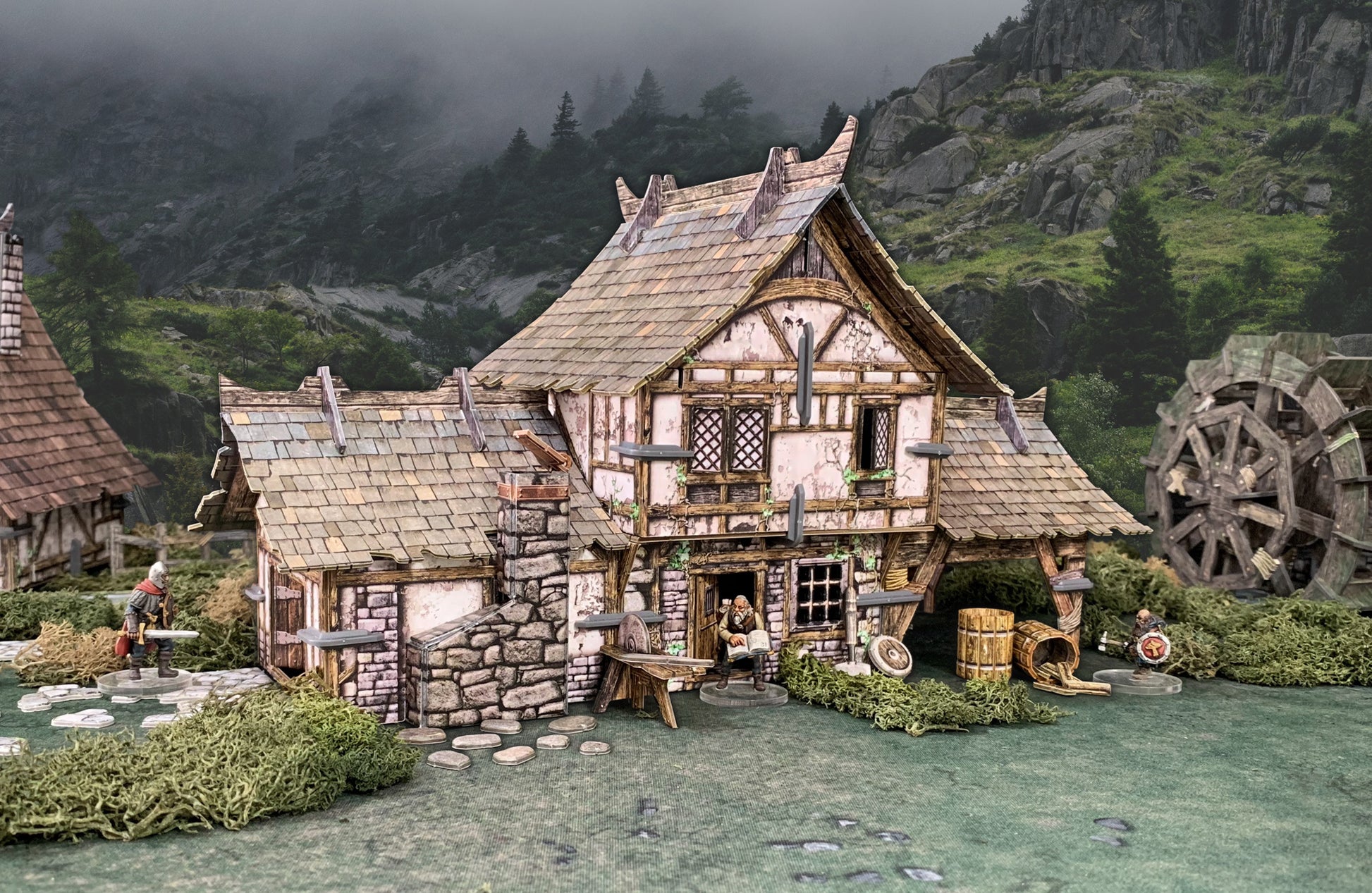 A detailed miniature fantasy village diorama featuring a timber-framed house, a reading figure, warriors, and a water wheel.
