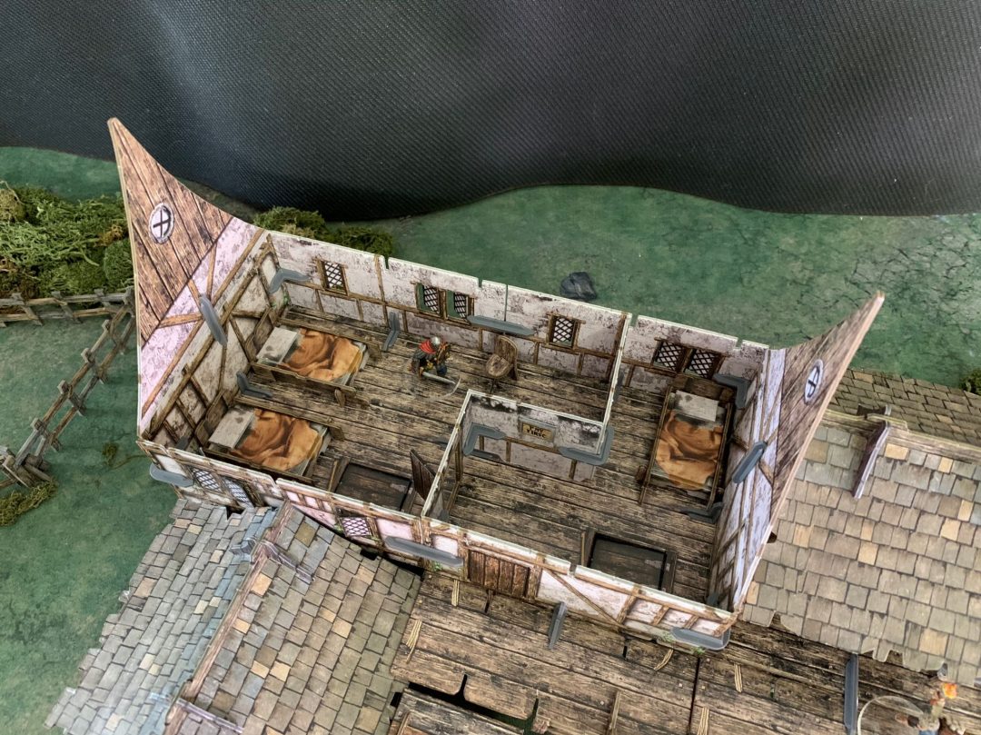 Overhead view of a miniature tabletop inn interior with wooden floors, beds, and a warrior figure standing inside.