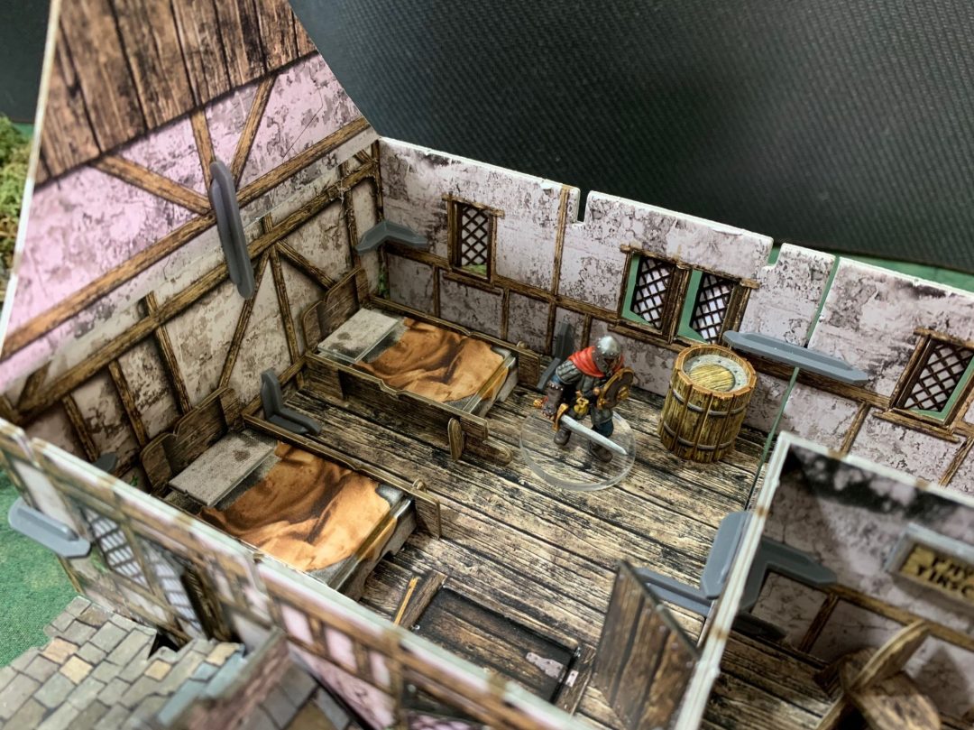 An armored miniature warrior with a sword and shield stands in a detailed tabletop game room with beds and a barrel.