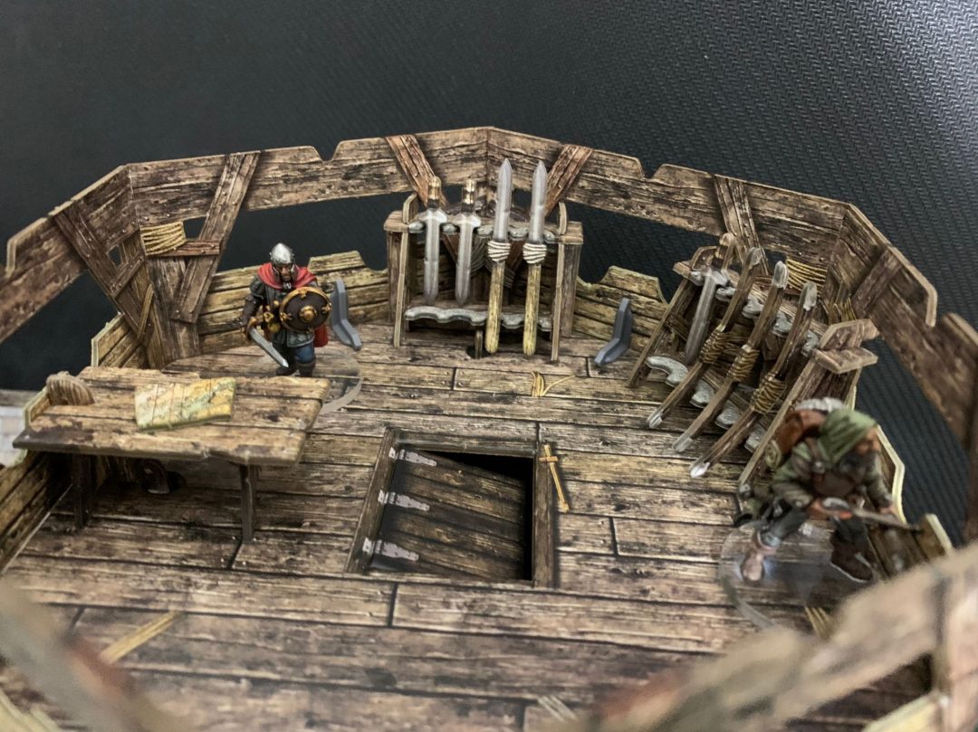 Two fantasy miniatures: a knight and a crouching ranger on a wooden ship's deck, surrounded by weapons, a map table, and an open trapdoor.