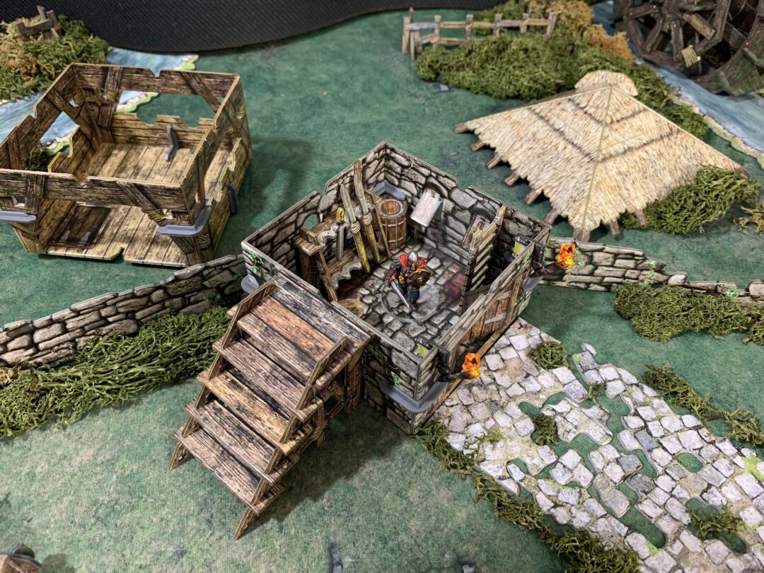 A miniature knight stands in a stone RPG building with stairs, surrounded by ruins, a thatched hut, and small flames.