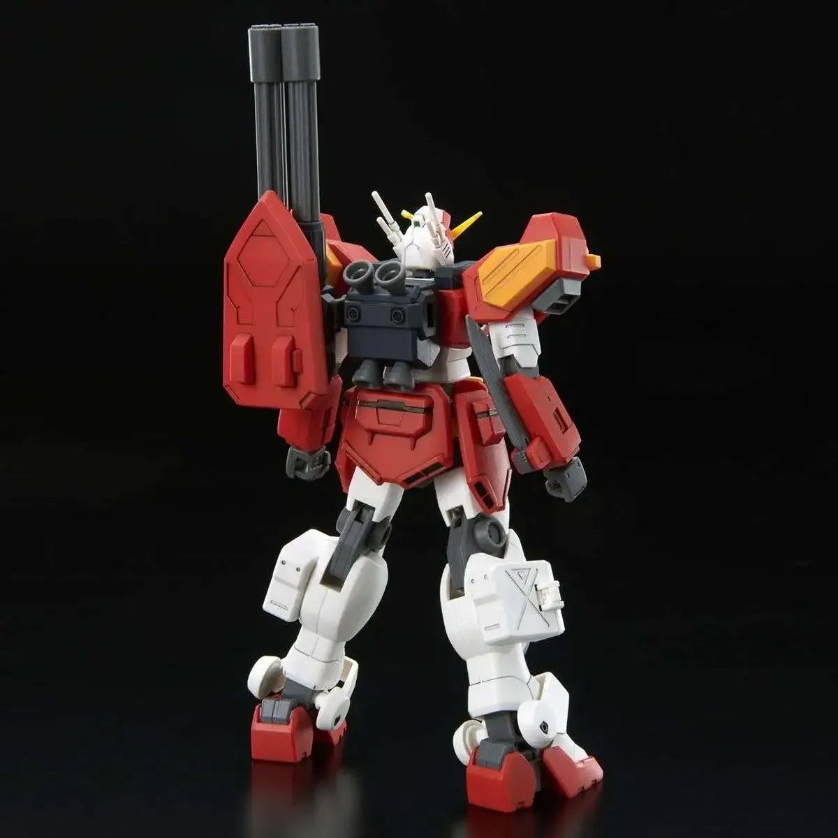 A red and white Gundam model stands with a large shoulder cannon and a shield, facing slightly away