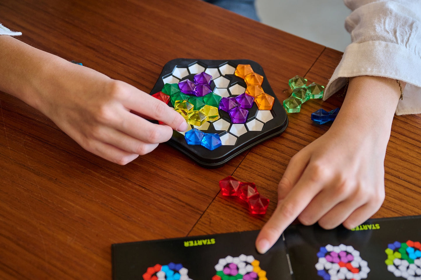 Overhead view of hands playing a colorful logic puzzle game with gem-like pieces on a wooden table