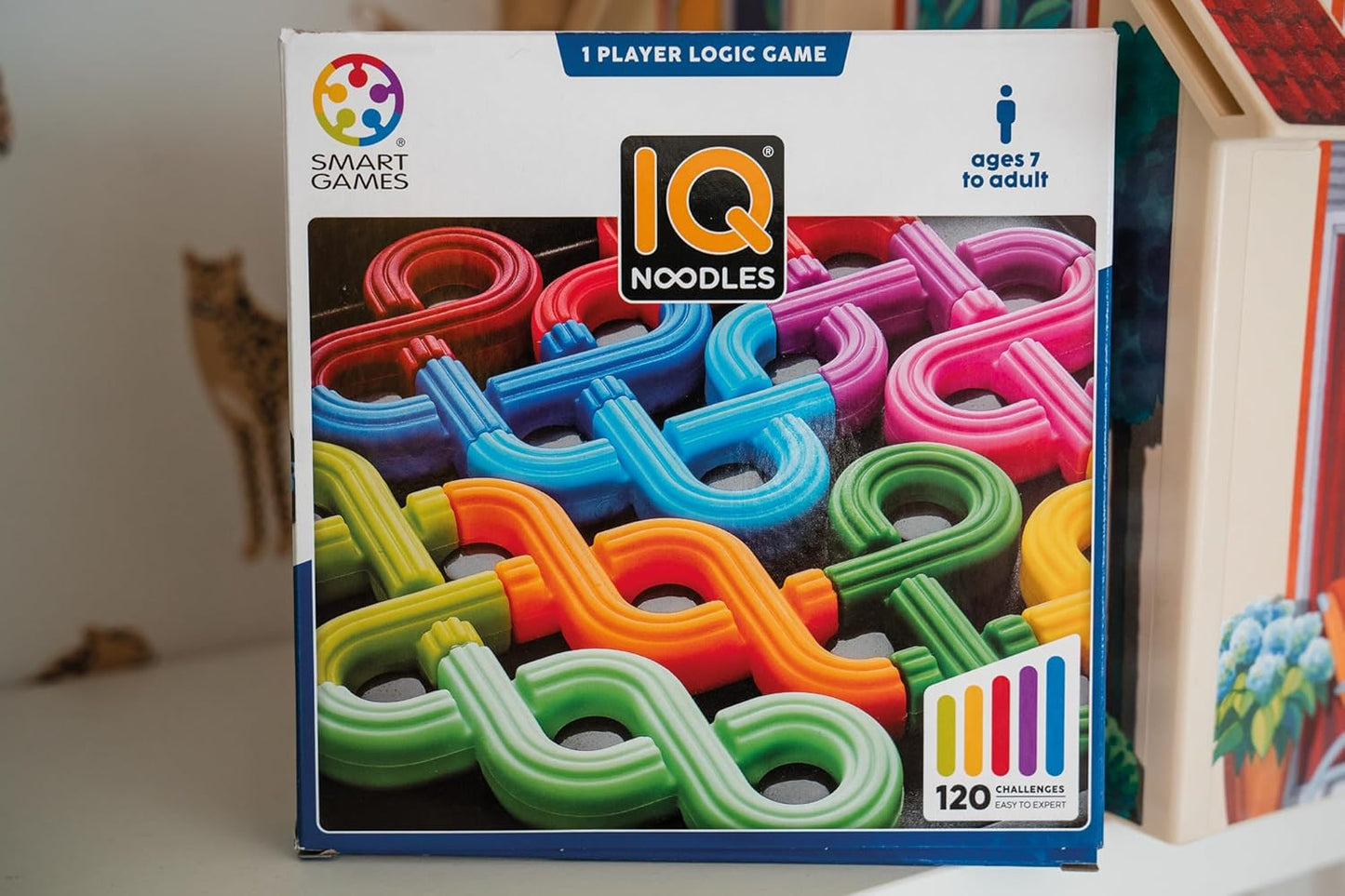 IQ Noodles logic game box showing colorful noodle-shaped pieces. Text: "1 player, ages 7 to adult"