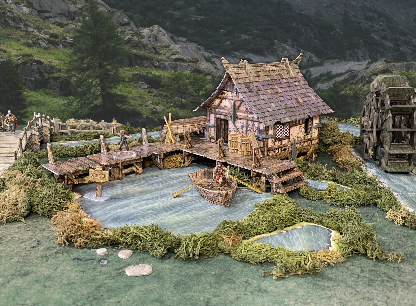 A detailed tabletop fantasy diorama of a riverside house with a water wheel, wooden dock, boat, and miniature adventurers.
