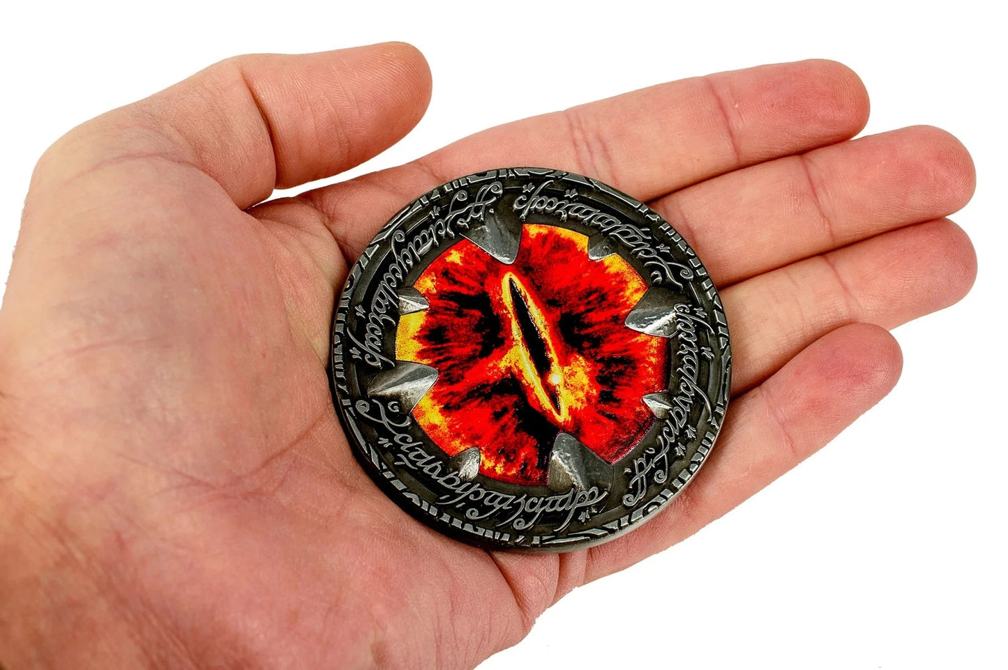 Hand holds a metal medallion featuring the fiery red Eye of Sauron and engraved script border