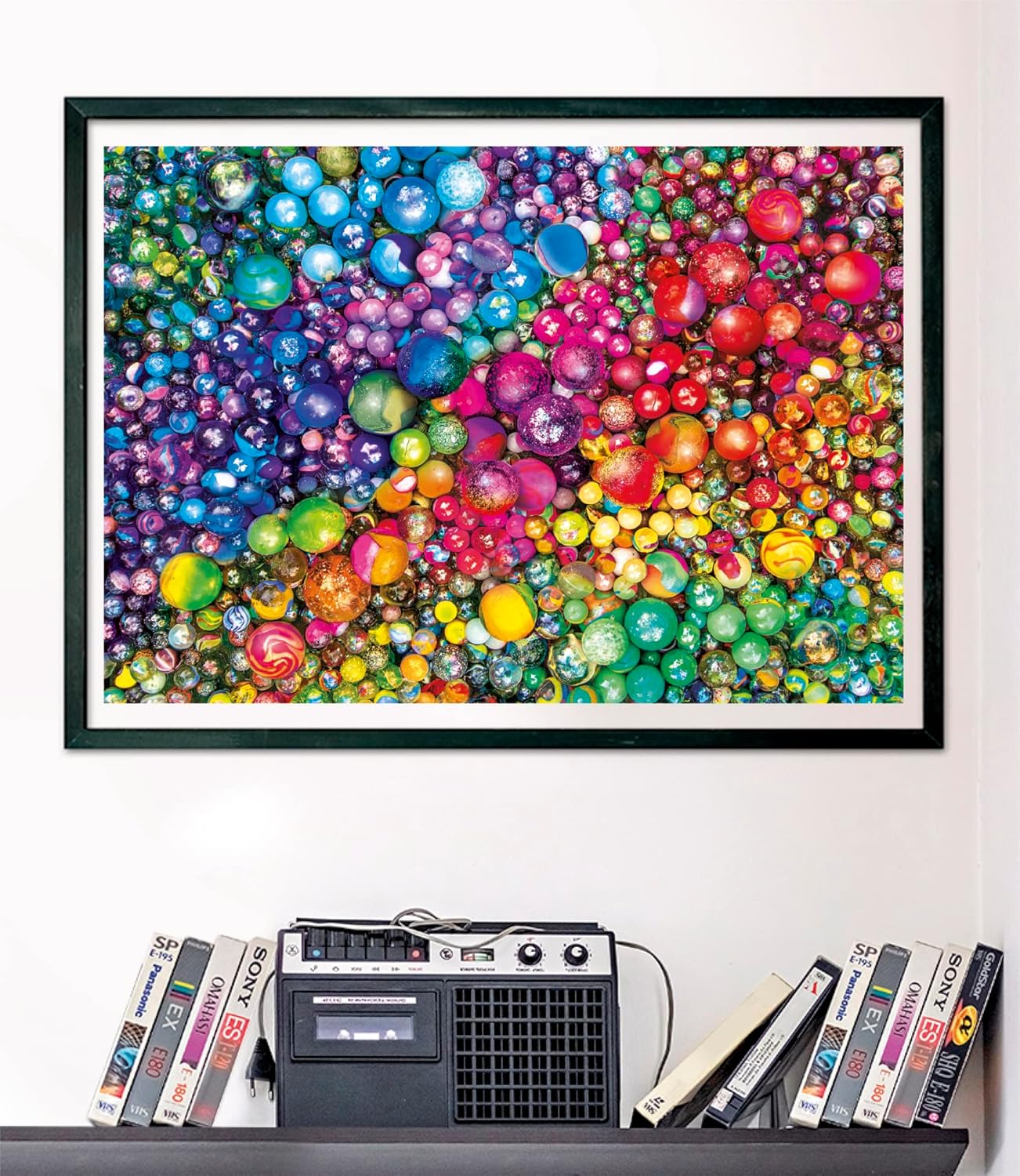 A framed vibrant artwork filled with countless colorful, shiny marbles in a rainbow pattern, displayed above a retro boombox and VHS tapes