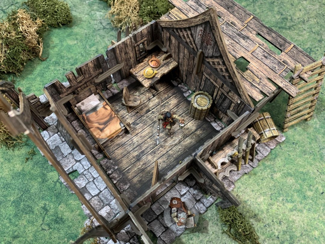 A miniature weathered wooden house interior for an RPG, featuring a character, bed, table with bowls, and barrels on a green mat.