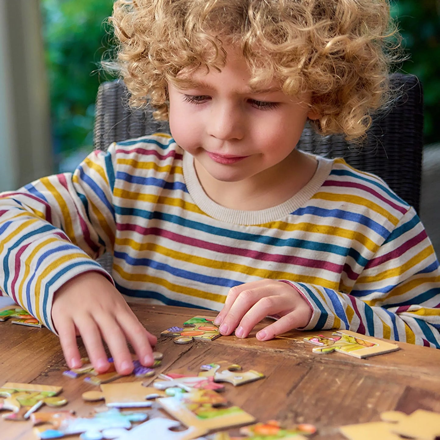Young boy with curly blonde hair working intently on a jigsaw puzzle at a wooden table