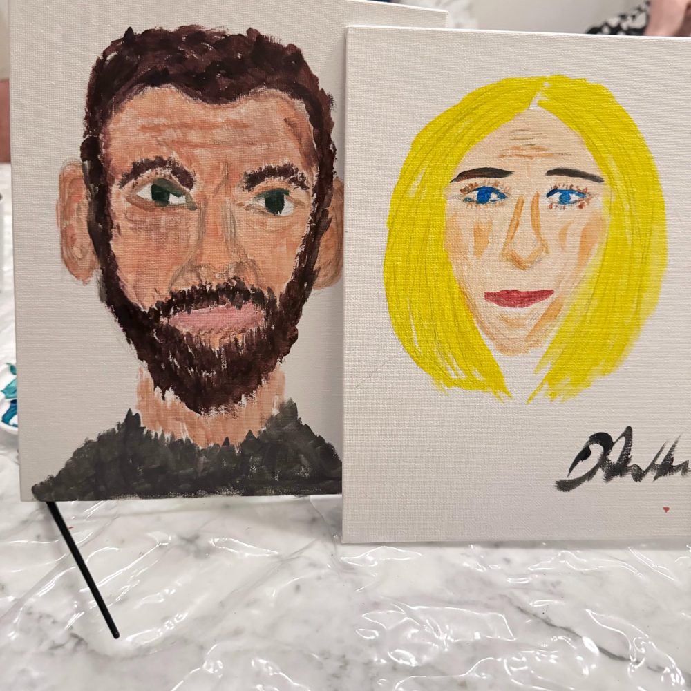 Two expressive painted portraits on small canvases: a bearded man and a yellow-haired woman, displayed on a plastic-covered surface