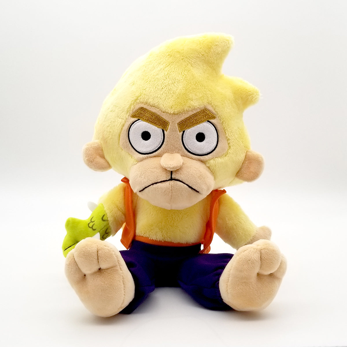Yellow monkey-like plush toy with a grumpy expression, orange suspenders, purple pants, and a green scaled arm sitting on a white background
