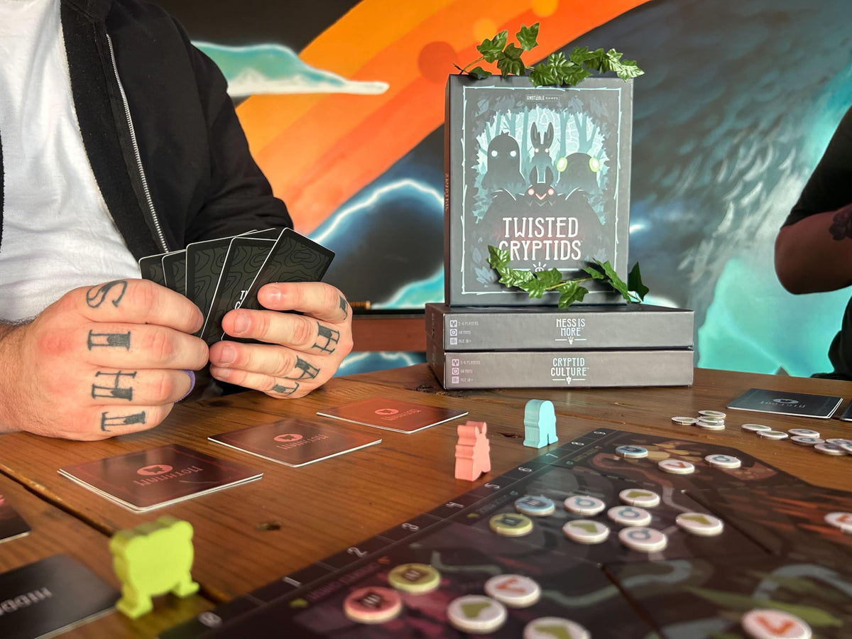 A person plays the Twisted Cryptids board game, holding cards while game pieces, tokens, and game boxes, including the Cryptid Culture expansion, are on a wooden table
