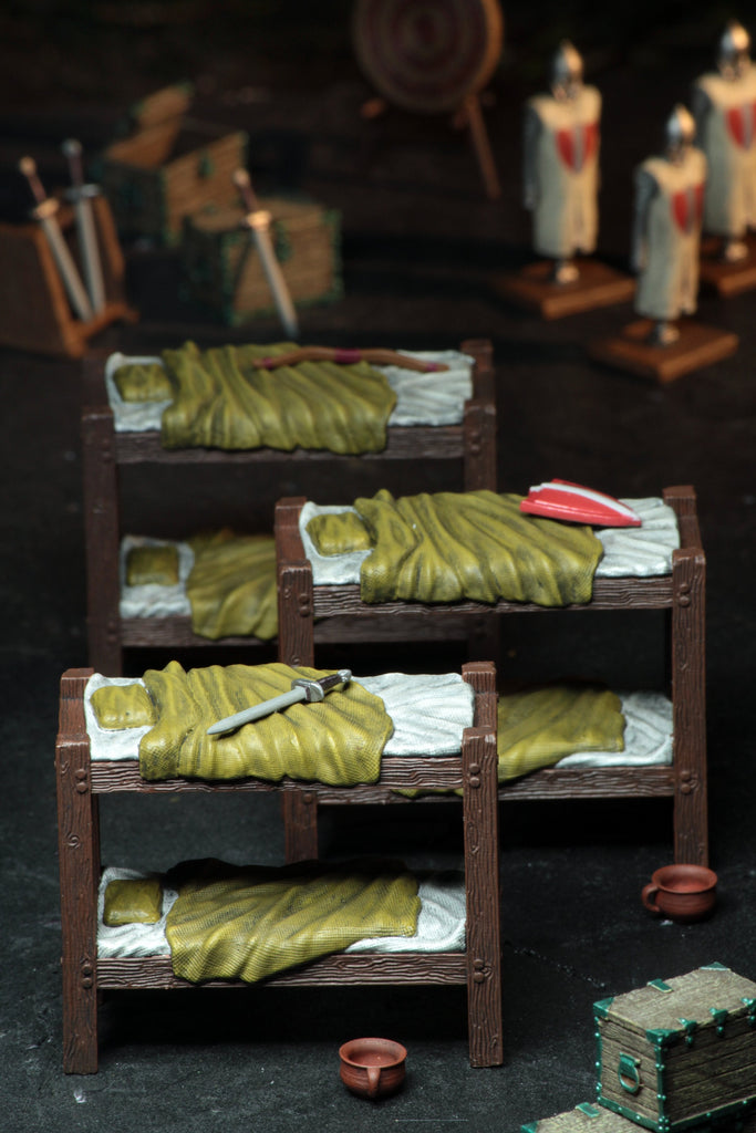 Miniature medieval barracks scene: three bunk beds with green blankets, a sword, and a shield. Blurred knights and chests behind