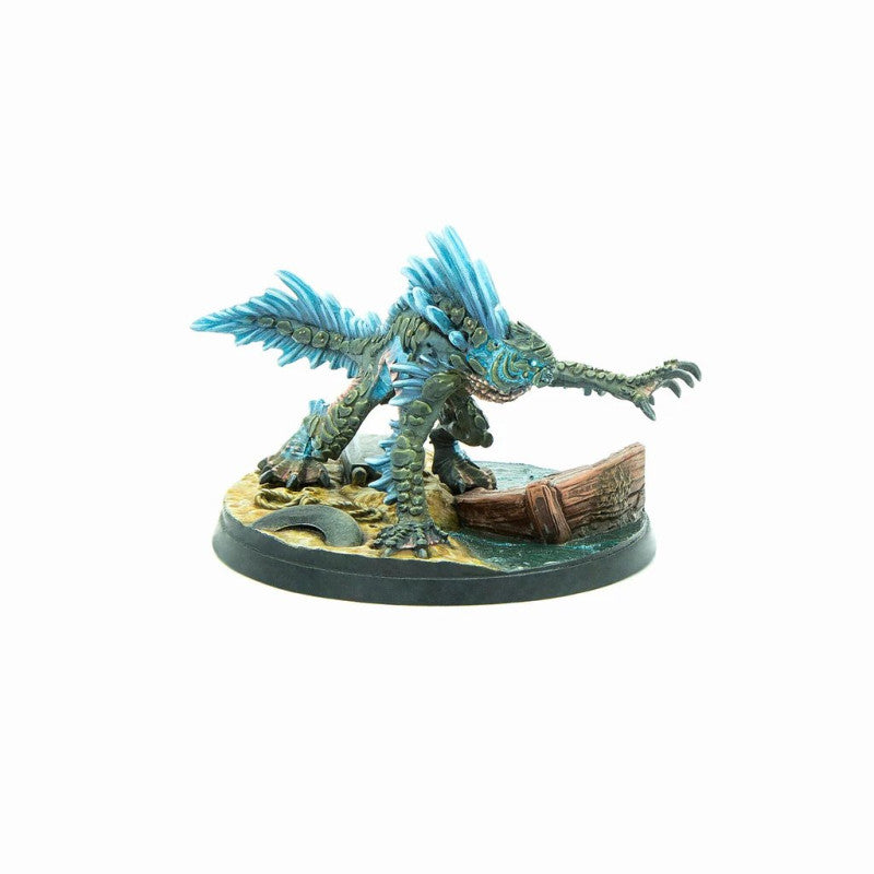 Miniature of a green, scaled aquatic monster with blue fin-like spines, crouching on a shore with a boat wreck.