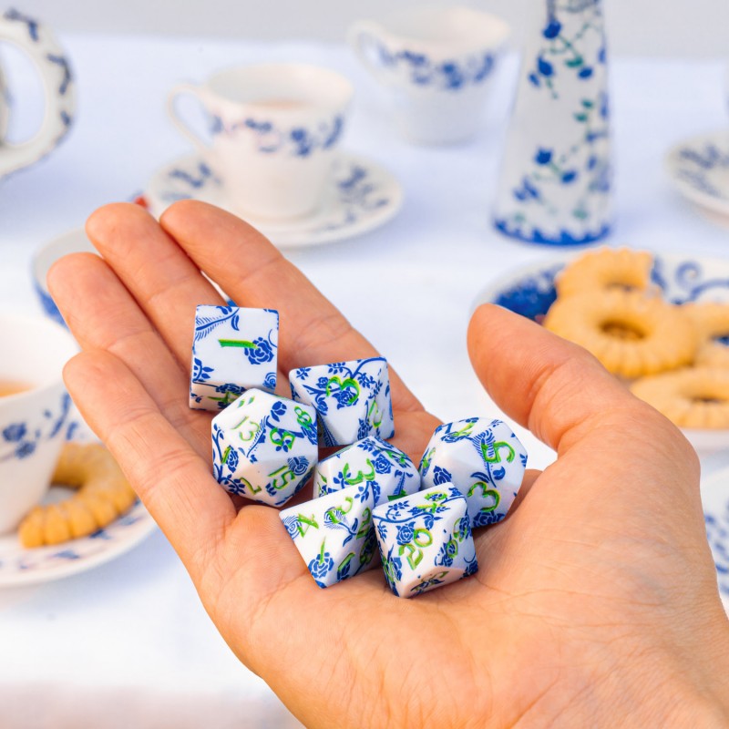 A hand holds a Full Art Porcelain RPG Dice Set, featuring white dice with blue and green floral patterns, against a background of matching tea cups and cookies