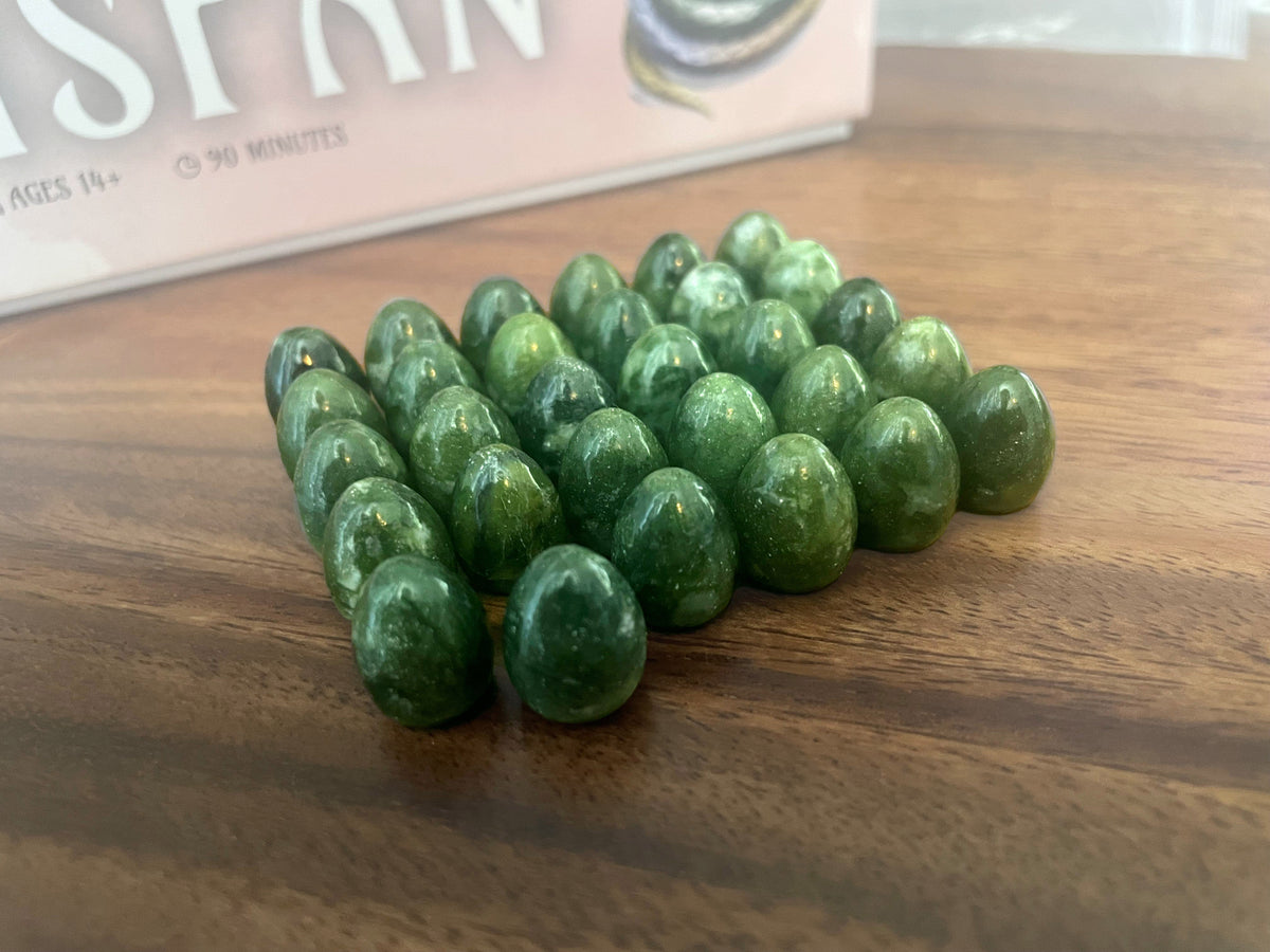 Polished green, egg-shaped game tokens clustered on a wooden table, near a blurred board game box