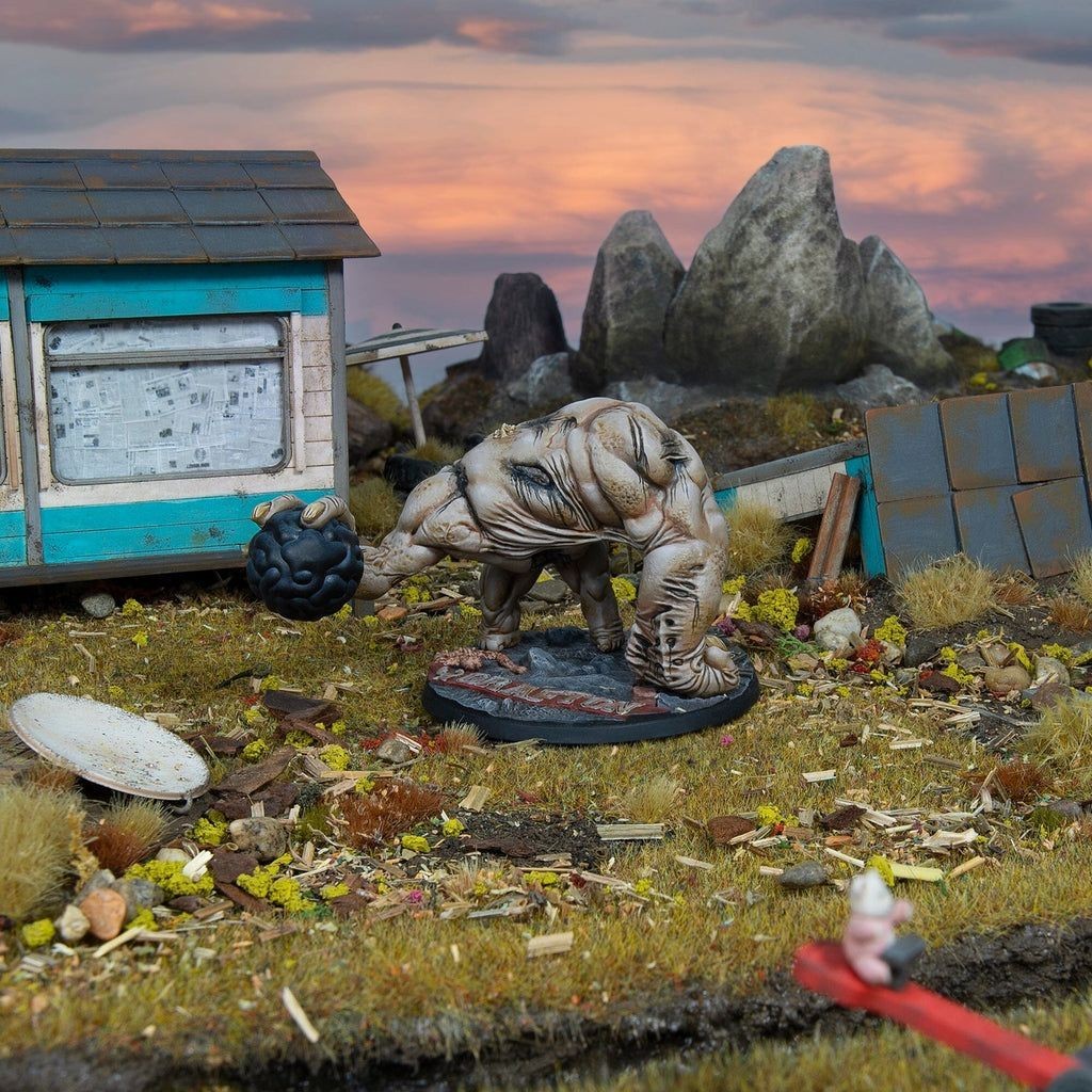 Hulking horror miniature holding a black orb in a derelict diorama with a blue shack and rocky backdrop at sunset
