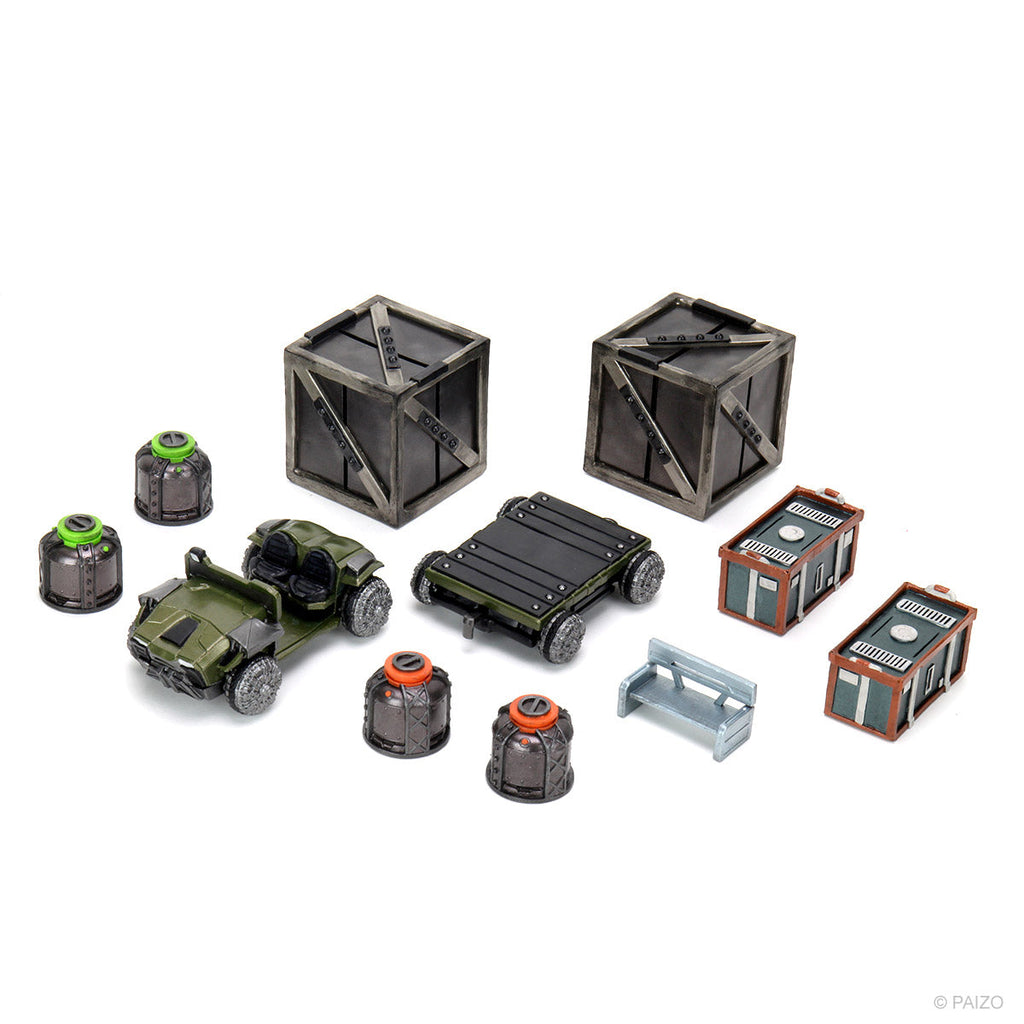 A collection of miniature game terrain: two crates, a green buggy, a flat cart, four canisters, two small buildings, and a bench