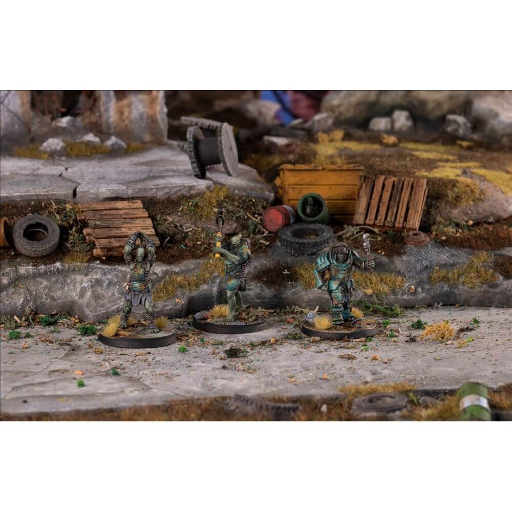 Three ghoulish miniatures with axes and weapons stand on a textured post-apocalyptic diorama with rubble