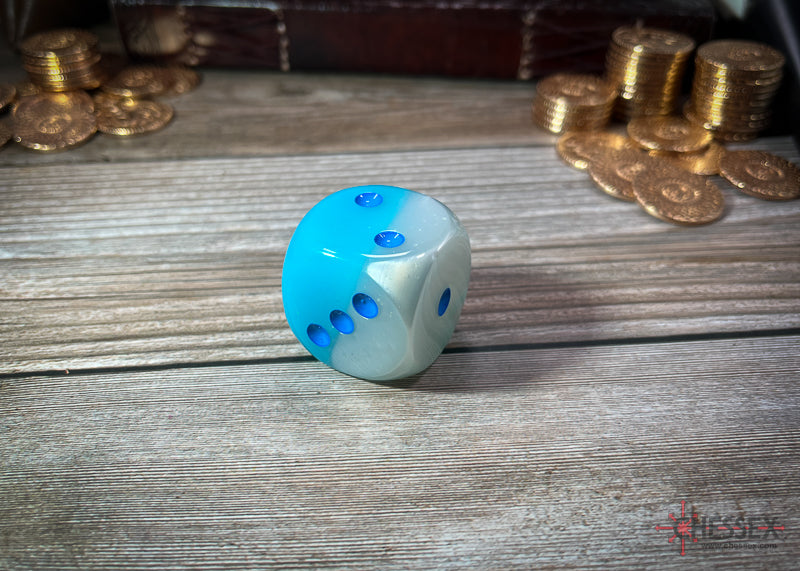 A Chessex Gemini two-toned blue and white dice with blue pips rests on a wooden table with blurred gold coins