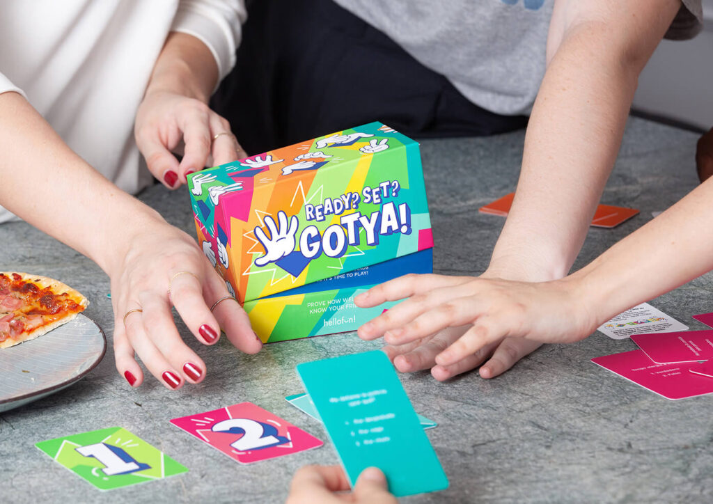 Hands playing a colorful "Ready? Set? Gotya!" card game with numbered cards on a table, a pizza slice is nearby