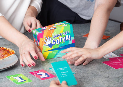 Hands playing a colorful "Ready? Set? Gotya!" card game with numbered cards on a table, a pizza slice is nearby