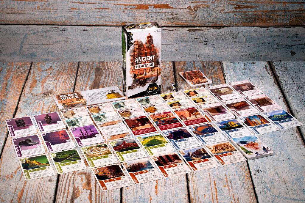 Ancient Knowledge Heritage board game box and numerous game cards displayed on a light blue wooden table showing intricate illustrations and game elements
