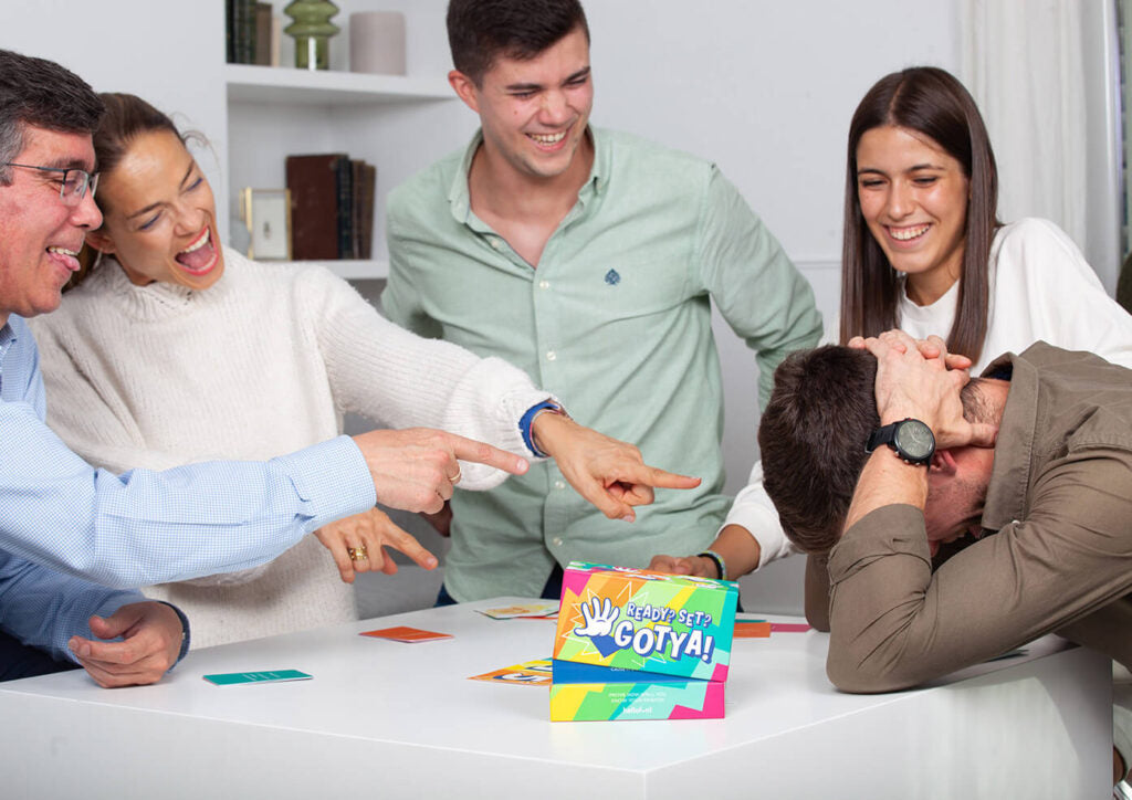 A group of friends laugh, point, and play the "Ready? Set? Gotya!" board game; one man covers his face