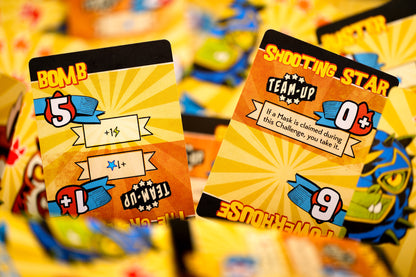 Two colorful board game cards, 'BOMB' and 'SHOOTING STAR,' with sunburst backgrounds, text, and numbers, on a pile of cards.