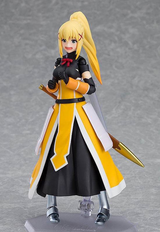 A Good Smile Figma Darkness figure stands smiling, showcasing her blond ponytail, red bow, and iconic yellow and black armor with a spear on her back